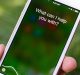 A hand holding an iPhone with the screen displaying the message "What can I help you with?" indicates that the voice assistant feature is active. In the background, blurred greenery hints that Siri shortcuts could help you manage your fitness and health routines efficiently.