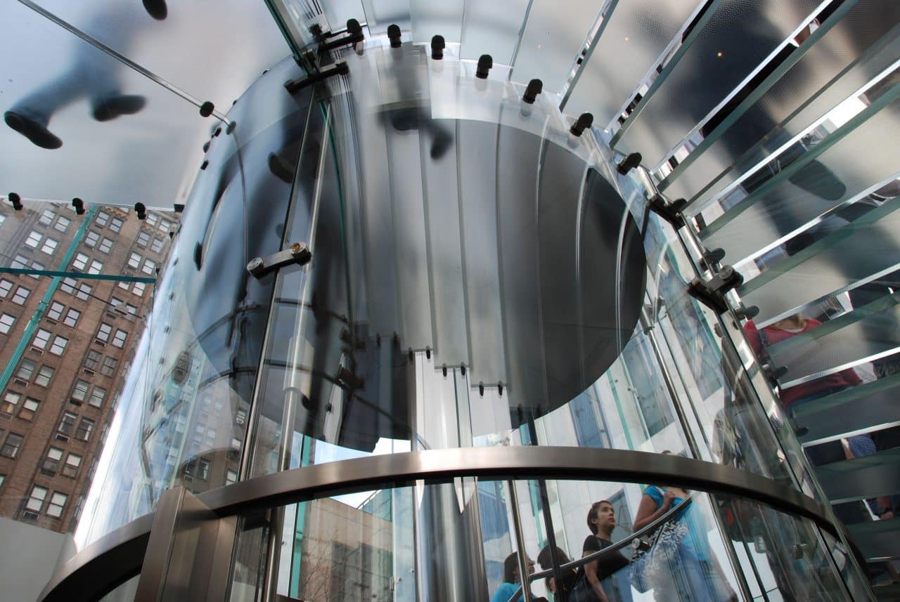 Spiral glass staircase inside a modern building, with team members walking up and down. The ceiling is made of glass, revealing tall buildings outside. The structure showcases sleek, contemporary design with metal and glass elements, inspiring anyone to join the team and advance their career.