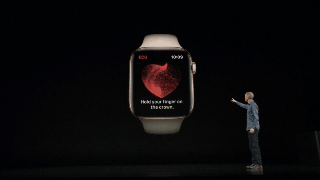 A person stands on a dark stage, pointing towards a large projected image of an Apple Watch displaying an ECG heart reading with the instruction, "Hold your finger on the crown." The time on the watch reads 10:09.