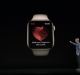 A person stands on a dark stage, pointing towards a large projected image of an Apple Watch displaying an ECG heart reading with the instruction, "Hold your finger on the crown." The time on the watch reads 10:09.