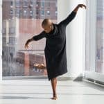 A person with a shaved head wearing a black dress performs a dance pose in a bright room with large windows. One leg is raised behind, and arms are outstretched gracefully. Sunlight streams in through the windows, illuminating the space and cityscape outside as they wear AirPods 2.