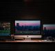 A dimly lit workspace featuring a 2019 Apple iMac setup with three monitors, each displaying a cityscape at dusk. The desk holds a keyboard, mouse, headphones, a notepad, pens, and two speakers. Various editing software is open on the screens.