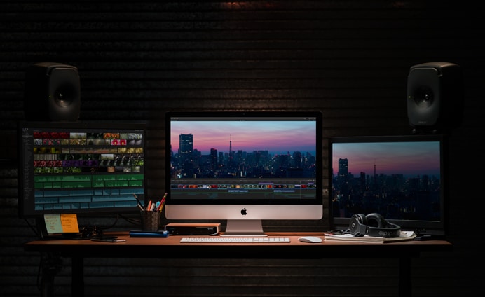 A dimly lit workspace featuring a 2019 Apple iMac setup with three monitors, each displaying a cityscape at dusk. The desk holds a keyboard, mouse, headphones, a notepad, pens, and two speakers. Various editing software is open on the screens.