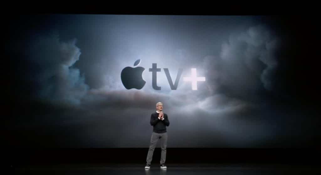 A person in a black sweater, gray pants, and white sneakers stands on a dark stage with a large screen behind them displaying the Apple TV+ logo against a cloudy background, highlighting the new Apple TV+ app.