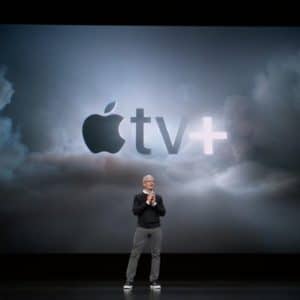 A person in a black sweater, gray pants, and white sneakers stands on a dark stage with a large screen behind them displaying the Apple TV+ logo against a cloudy background, highlighting the new Apple TV+ app.