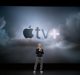 A person in a black sweater, gray pants, and white sneakers stands on a dark stage with a large screen behind them displaying the Apple TV+ logo against a cloudy background, highlighting the new Apple TV+ app.