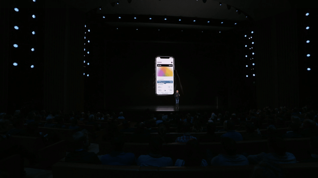 An individual stands on a dark stage with spotlights at the front, addressing a seated audience. Behind the person, a large screen displays a smartphone showcasing an app interface with gradient colors and prominently featuring the new Apple Card credit card.