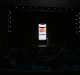 An individual stands on a dark stage with spotlights at the front, addressing a seated audience. Behind the person, a large screen displays a smartphone showcasing an app interface with gradient colors and prominently featuring the new Apple Card credit card.