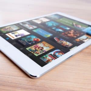 A white low-cost iPad lies on a wooden surface, displaying a grid of colorful movie and game icons on its screen, including titles like Toy Story and Cars. The tablet is tilted at a slight angle, highlighting its thin profile and fine details.