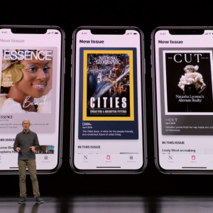 A person stands on stage in front of a large screen displaying three smartphones. Each phone shows the cover of a different magazine: "ESSENCE," "CITIES," and "CUT." The heading above each phone reads "New Issue" via Apple News+ for just $9.99/mo.