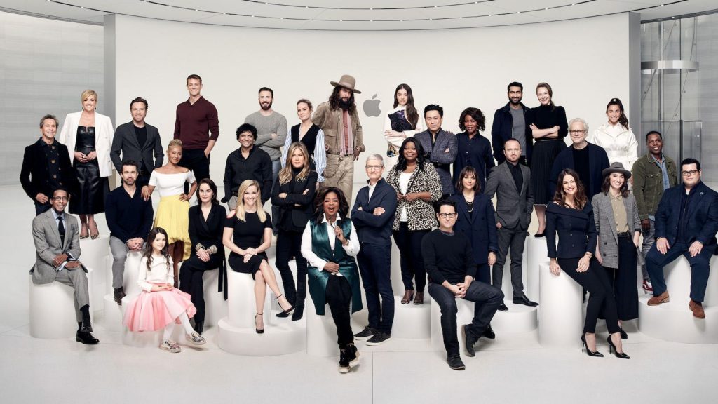 A group of diverse people, including men and women, pose together. Some are standing while others are seated on white steps and platforms. They are in a modern setting with a minimalist design, an Apple logo visible in the background like an unanswered question in a sleek advertisement.