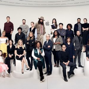 A group of diverse people, including men and women, pose together. Some are standing while others are seated on white steps and platforms. They are in a modern setting with a minimalist design, an Apple logo visible in the background like an unanswered question in a sleek advertisement.