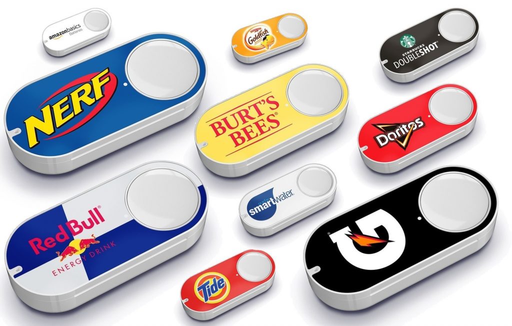 Several Amazon Dash Buttons are displayed, each branded with different logos, including Nerf, Goldfish, Burt's Bees, Starbucks Doubleshot, RedBull, Smartwater, Doritos, Tide, and Gatorade. The buttons are white with a large logo on each—a nostalgic reminder as Amazon discontinues the Dash Button line.