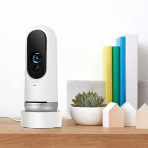 A modern white and black Lighthouse AI security camera sits on a wooden table. Next to it, there's a small potted succulent, a few colorful books standing upright, and two small house-shaped wooden decorations.