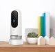 A modern white and black Lighthouse AI security camera sits on a wooden table. Next to it, there's a small potted succulent, a few colorful books standing upright, and two small house-shaped wooden decorations.