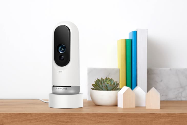 A modern white and black Lighthouse AI security camera sits on a wooden table. Next to it, there's a small potted succulent, a few colorful books standing upright, and two small house-shaped wooden decorations.