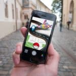 A person holds a smartphone outdoors on a cobblestone street, immersed in an augmented reality game featuring a Poké Ball, a path, and a creature. Historic-style buildings line the street, blending the charm of old-world architecture with modern AR technology.