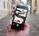 A person holds a smartphone outdoors on a cobblestone street, immersed in an augmented reality game featuring a Poké Ball, a path, and a creature. Historic-style buildings line the street, blending the charm of old-world architecture with modern AR technology.