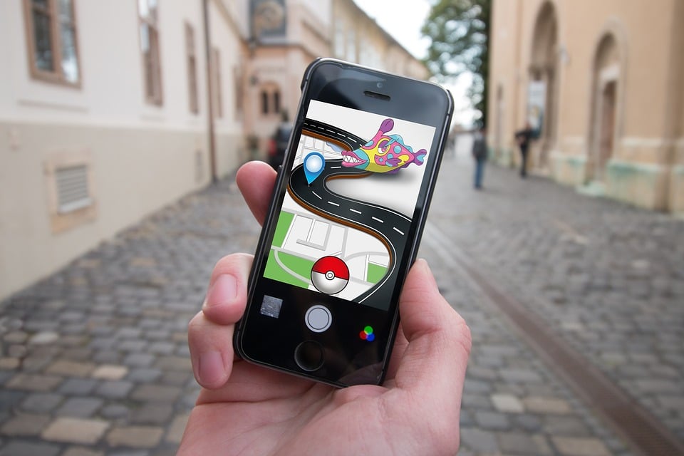 A person holds a smartphone outdoors on a cobblestone street, immersed in an augmented reality game featuring a Poké Ball, a path, and a creature. Historic-style buildings line the street, blending the charm of old-world architecture with modern AR technology.