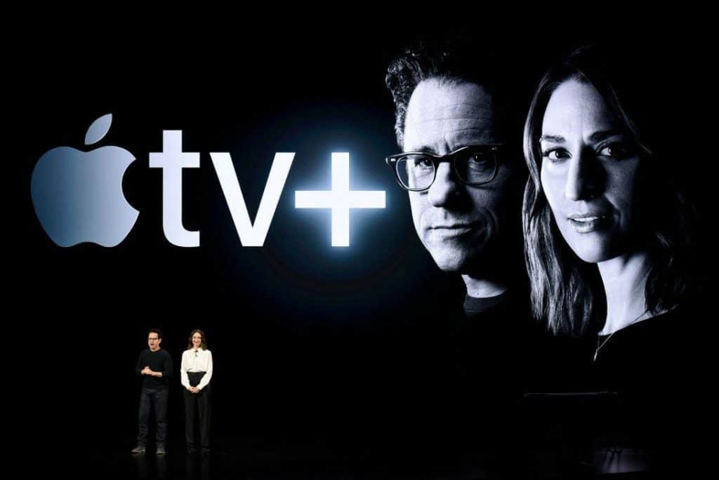 Two people stand on a dark stage in front of a large screen displaying the Apple TV+ logo. The screen also shows large black-and-white portraits of Stephen King and J.J. Abrams. The individuals on stage are facing the audience.