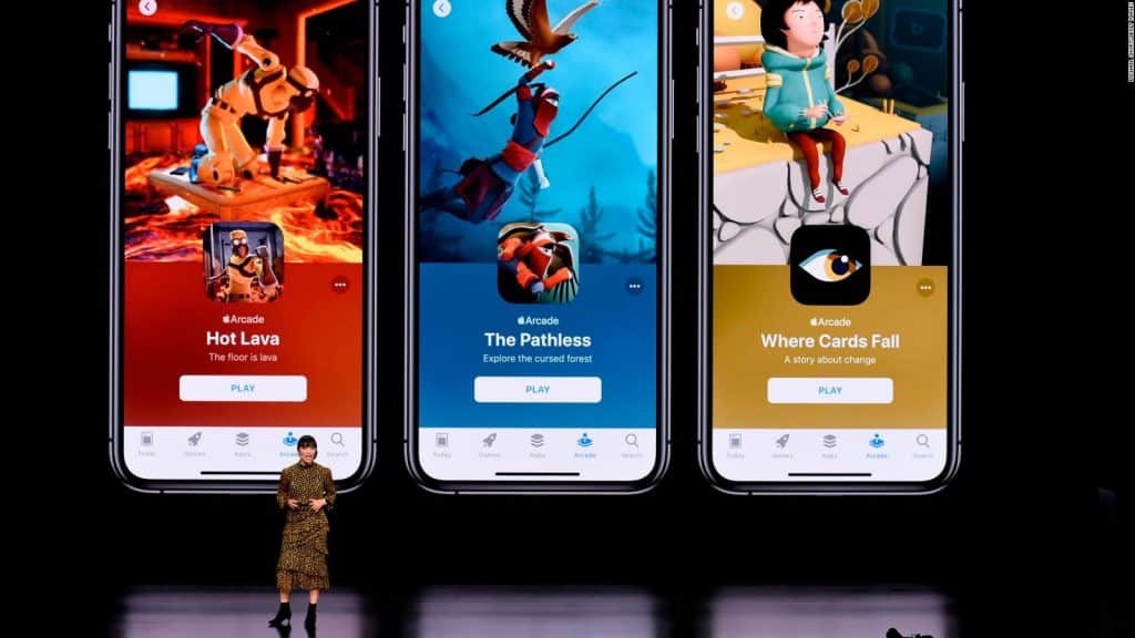 A person stands on stage giving a presentation in front of a large screen displaying three iPhone screens showcasing Arcade games: "Hot Lava," "The Pathless," and "Where Cards Fall." Gesturing with their right hand, they highlight Apple’s $500 million spending on this ambitious gaming service.