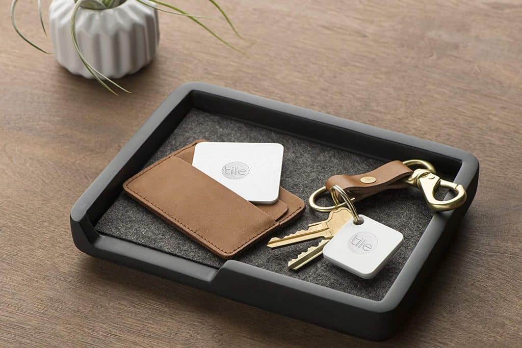 A neatly arranged tray on a wooden surface holds a small potted plant, a brown leather cardholder with a square Tile tracker, and a keychain with two keys and another square Tracking Tile attached.