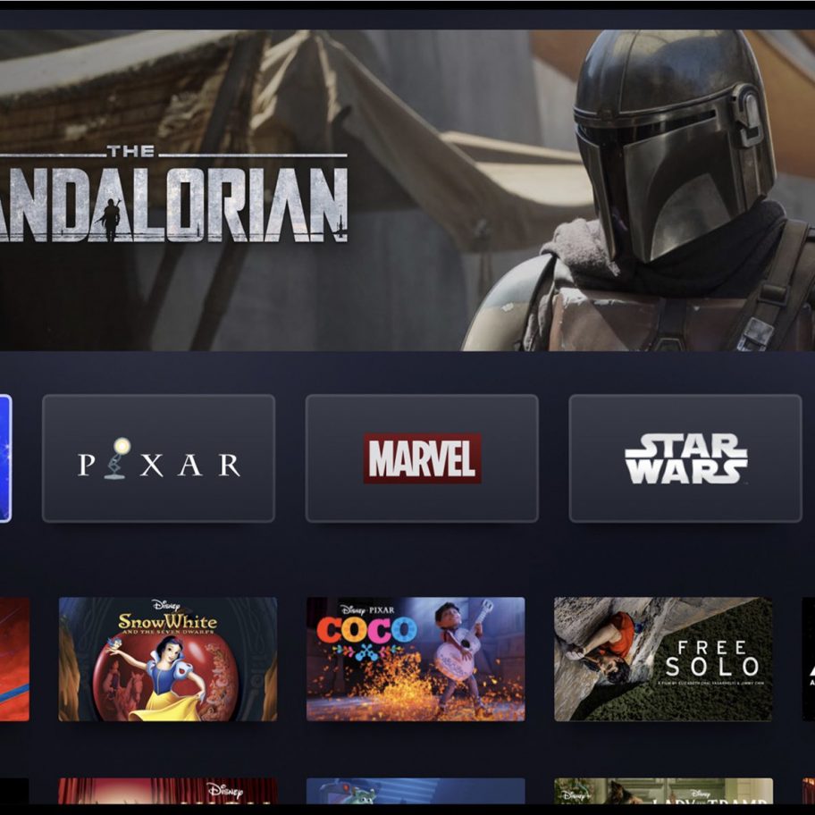 Screenshot of a streaming service interface showing various content options. Featured is "The Mandalorian" with a character in armor and helmet. Below are categories: Disney, Pixar, Marvel, Star Wars, National Geographic, and recommended titles like "Captain Marvel," demonstrating Bob Iger's vision as Disney CEO.