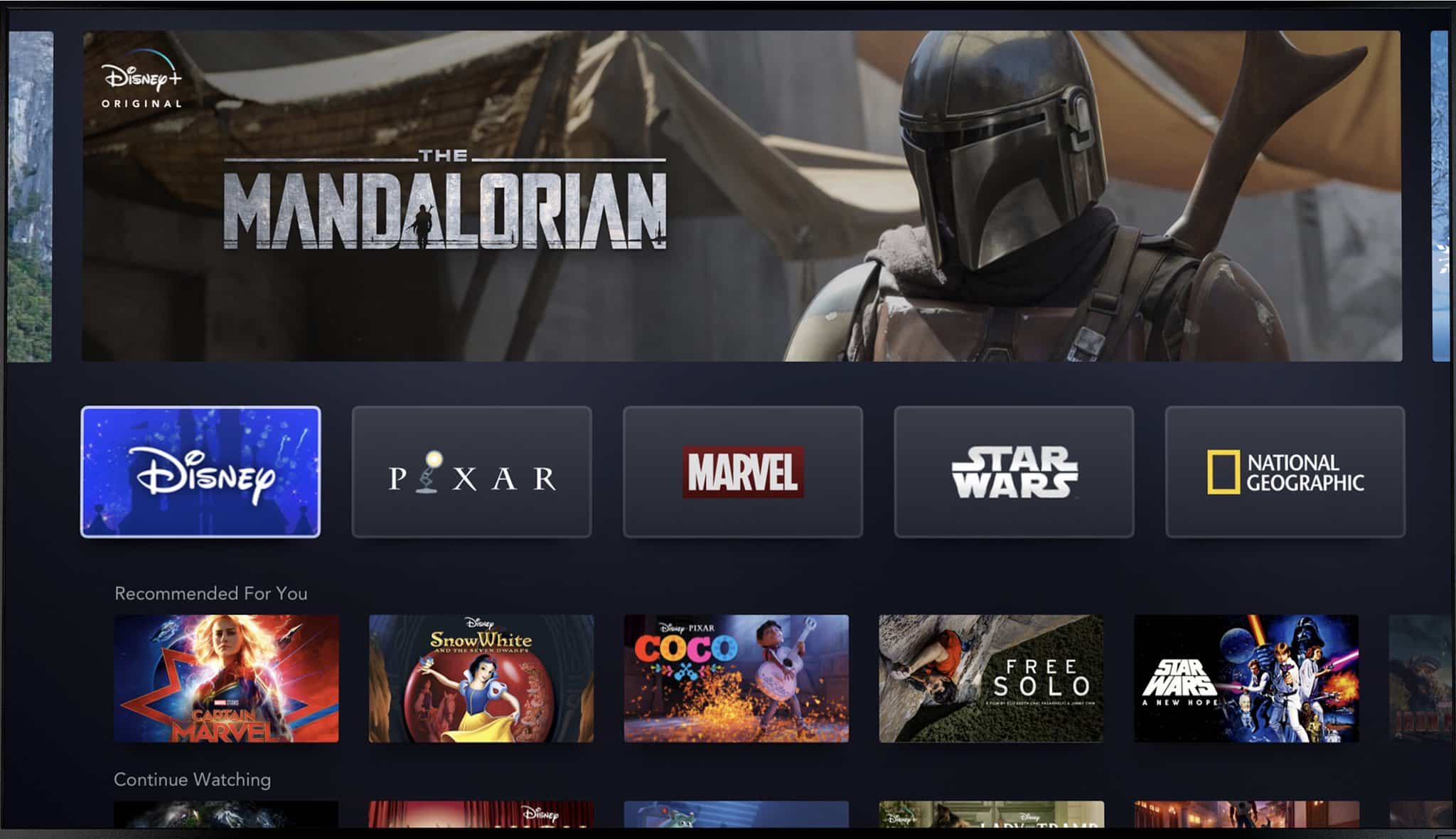 Screenshot of a streaming service interface showing various content options. Featured is "The Mandalorian" with a character in armor and helmet. Below are categories: Disney, Pixar, Marvel, Star Wars, National Geographic, and recommended titles like "Captain Marvel," demonstrating Bob Iger's vision as Disney CEO.