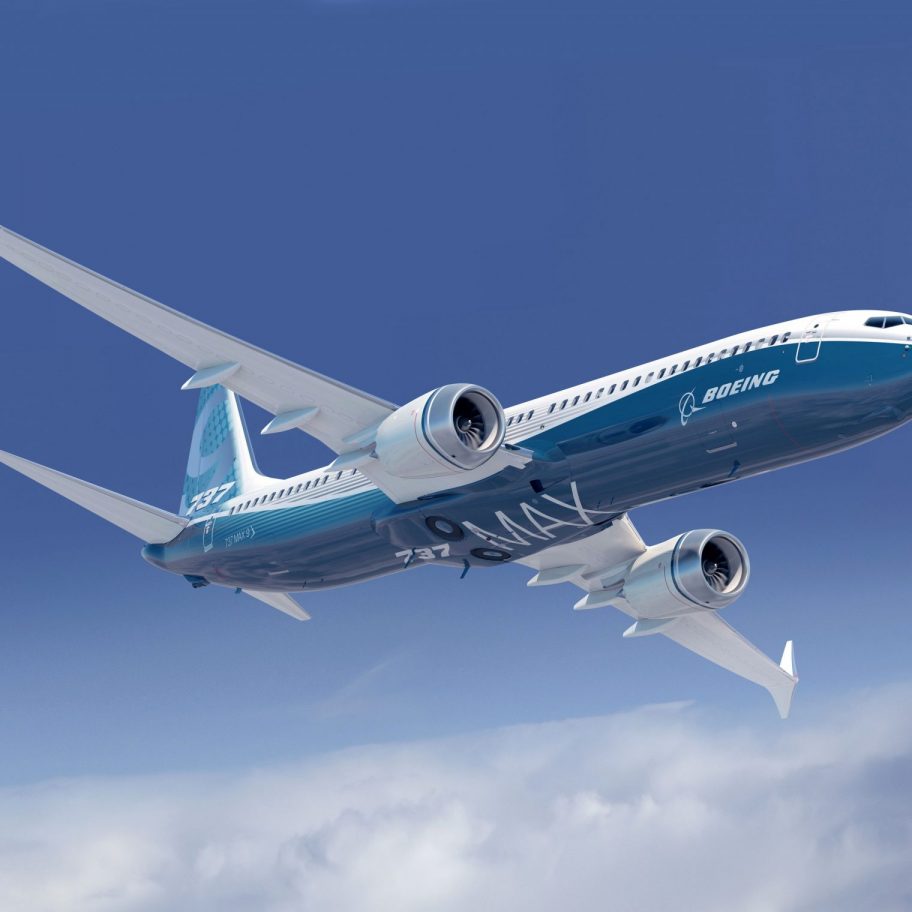 An airplane with blue and white livery marked "Boeing 737 MAX" ascends in a clear blue sky with a few clouds below. The aircraft, featuring automated systems and two engines mounted under its wings, boasts a sleek, modern design.
