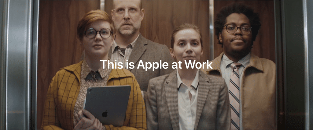 Four people stand in an elevator, facing forward. The group includes a woman with short red hair holding an iPad, a man in glasses and a suit, a woman in a beige blazer, and a man with a beard and glasses wearing a pink tie. Text on the image reads "This is Apple at Work," highlighting its appeal to business customers.