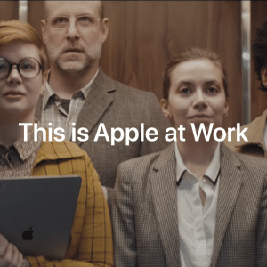 Four people stand in an elevator, facing forward. The group includes a woman with short red hair holding an iPad, a man in glasses and a suit, a woman in a beige blazer, and a man with a beard and glasses wearing a pink tie. Text on the image reads "This is Apple at Work," highlighting its appeal to business customers.