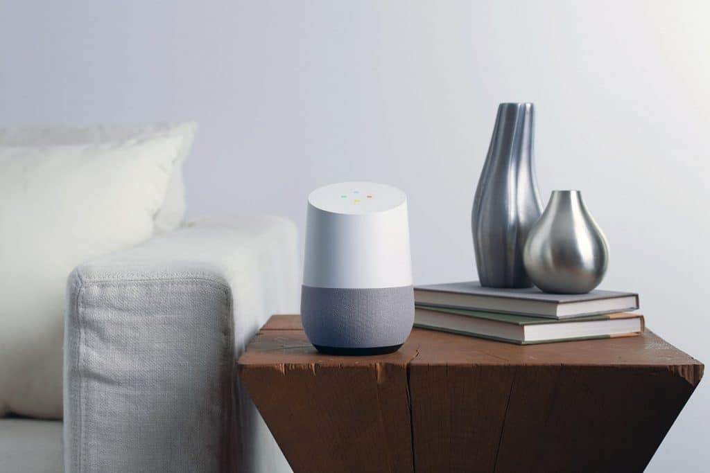 A smart speaker, possibly a Google device, sits on a wooden side table beside two metallic vases and several stacked books. The table is next to a light-colored sofa against a plain, white wall, giving the room a modern and minimalistic look. Ad-supported music fills the space with gentle ambiance.