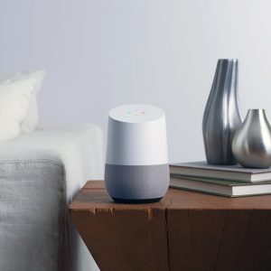 A smart speaker, possibly a Google device, sits on a wooden side table beside two metallic vases and several stacked books. The table is next to a light-colored sofa against a plain, white wall, giving the room a modern and minimalistic look. Ad-supported music fills the space with gentle ambiance.