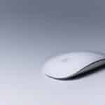 A sleek white wireless Apple Magic Mouse rests on a smooth, light grey surface. The mouse features a minimalist design with rounded edges and a glossy finish, displaying the Apple logo subtly at the center. With iPad Mouse Support in iOS 13, this Magic Mouse becomes even more versatile.