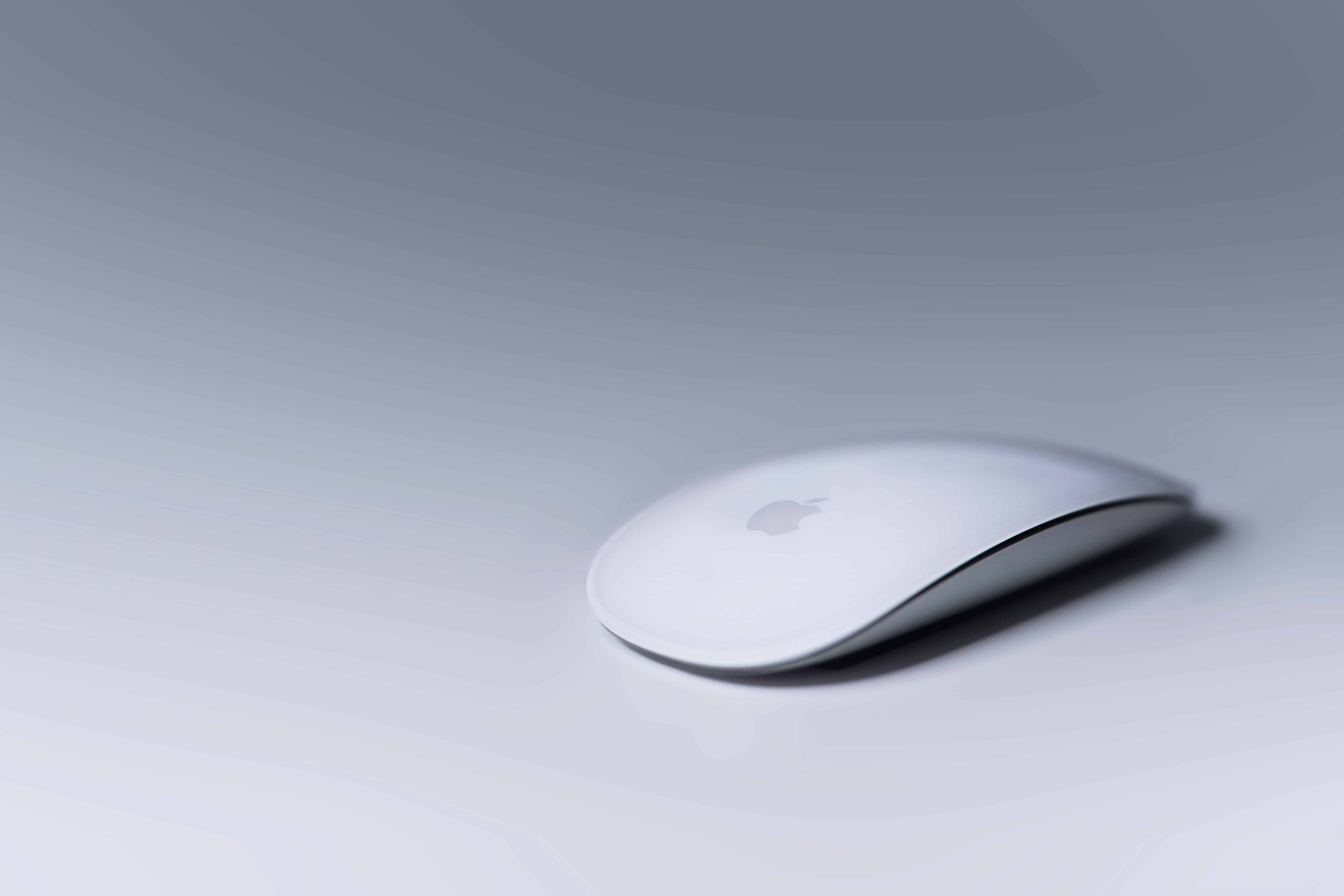 A sleek white wireless Apple Magic Mouse rests on a smooth, light grey surface. The mouse features a minimalist design with rounded edges and a glossy finish, displaying the Apple logo subtly at the center. With iPad Mouse Support in iOS 13, this Magic Mouse becomes even more versatile.