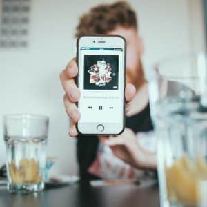 A US teen holds up an iPhone displaying a music player app, with album artwork and playback controls visible. In the background, there’s a blurred view of glasses with drinks, a wooden bowl, and a table setting.