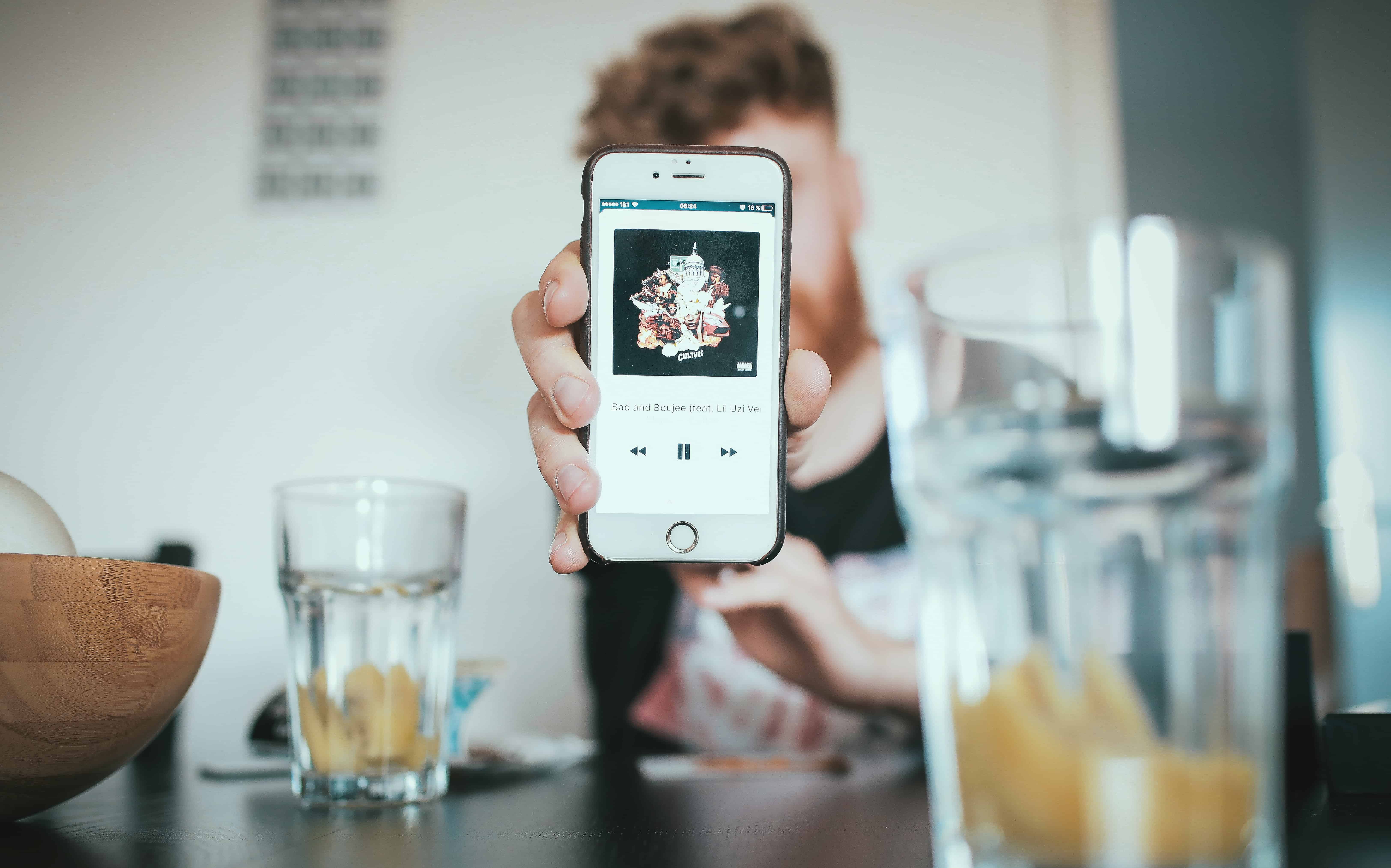 A US teen holds up an iPhone displaying a music player app, with album artwork and playback controls visible. In the background, there’s a blurred view of glasses with drinks, a wooden bowl, and a table setting.