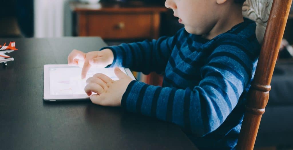 A young child, wearing a blue and black striped shirt, sits at a table and interacts with a tablet using both hands. An Apple logo is visible on the device. The background includes a wooden chair and a piece of furniture, partially in view, adding a cozy, homey atmosphere.