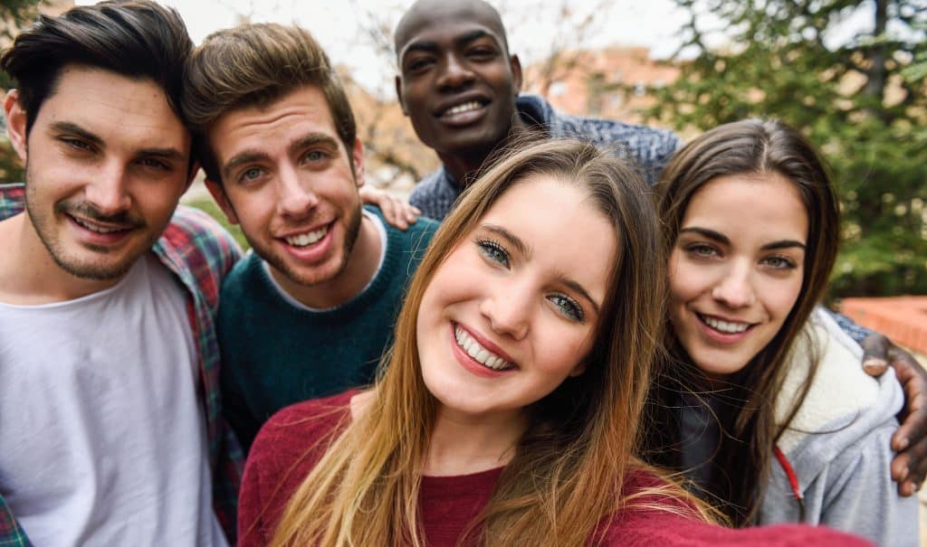 A group of five friends huddle close together outdoors, smiling brightly at the camera, seemingly oblivious to how social media can be damaging society. They appear to be enjoying a pleasant day, with trees and buildings visible in the blurred background. The friends' diverse expressions convey a sense of happiness and camaraderie.