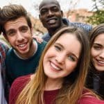 A group of five friends huddle close together outdoors, smiling brightly at the camera, seemingly oblivious to how social media can be damaging society. They appear to be enjoying a pleasant day, with trees and buildings visible in the blurred background. The friends' diverse expressions convey a sense of happiness and camaraderie.