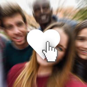 A blurred group selfie of five people with a large white heart icon and a pixelated cursor hand superimposed in the center, reminiscent of social media's "Like Me" culture. The background is out of focus, emphasizing the icon in the middle. The group appears to be outdoors.