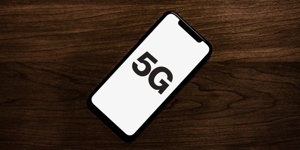 A sleek smartphone with a black frame lies on a dark wooden surface, displaying the text "5G" on its screen. This could be one of Apple’s 2020 5G iPhone models, part of the 200 million units shipped globally.