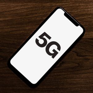 A sleek smartphone with a black frame lies on a dark wooden surface, displaying the text "5G" on its screen. This could be one of Apple’s 2020 5G iPhone models, part of the 200 million units shipped globally.