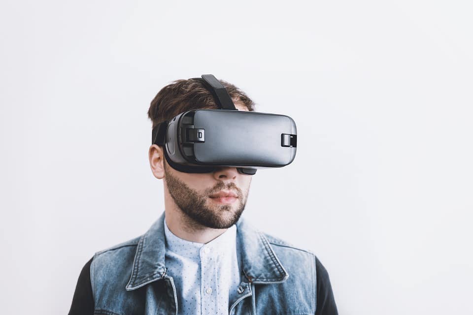 A bearded man wearing a virtual reality headset, with short dark hair and dressed in a white patterned shirt under a blue denim jacket, stands against a plain white background. This could easily be Arthur van Hoff engrossed in his latest VR research for Apple.