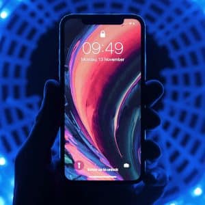 A hand holding a smartphone with a vibrant, abstract wallpaper displayed on the screen. The phone, likely one of the 2019 iPhones, shows the time as 09:49 and the date as Monday, 13 November. The background features a pattern of blue LED lights forming a circular tunnel effect.