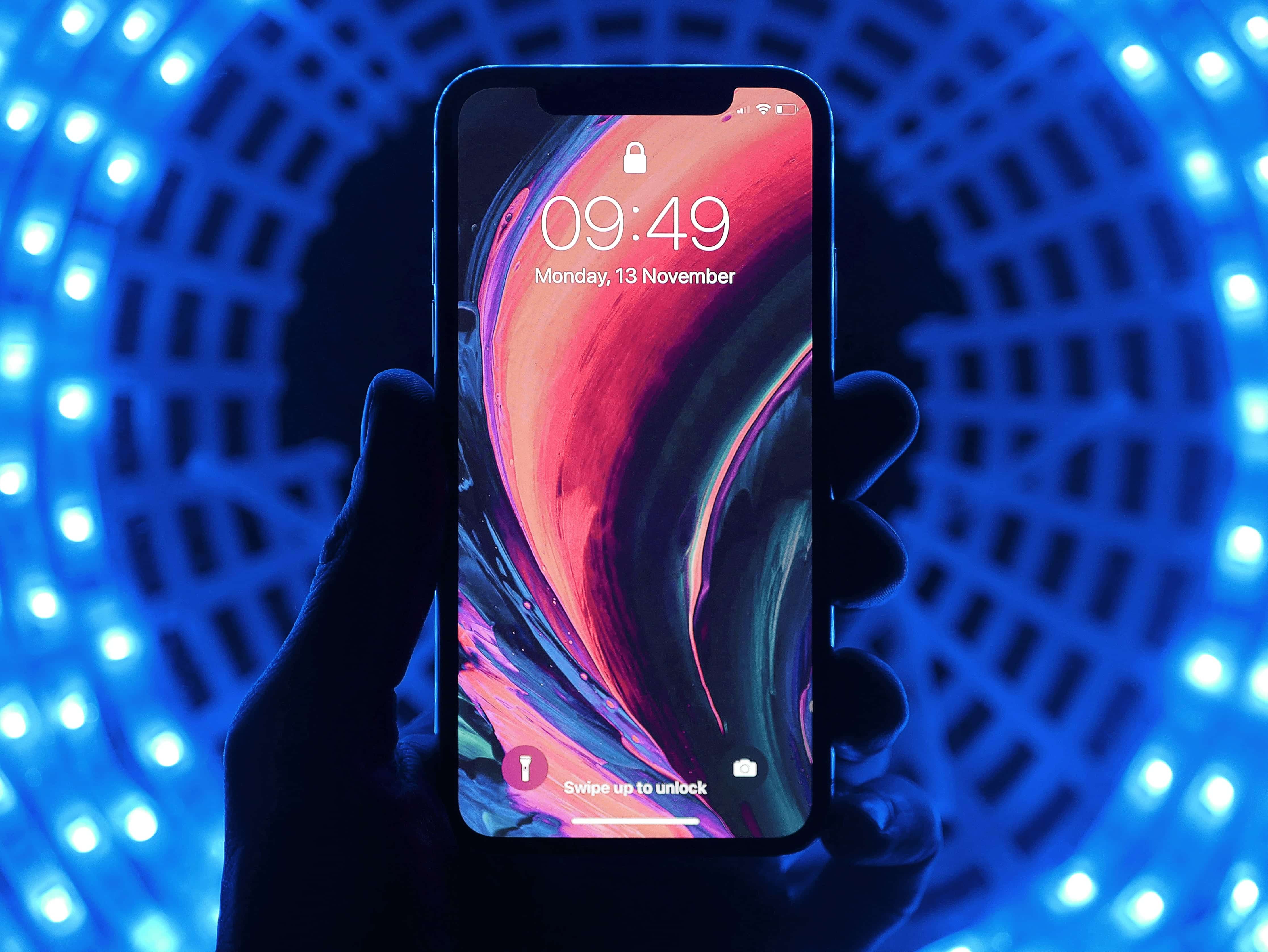 A hand holding a smartphone with a vibrant, abstract wallpaper displayed on the screen. The phone, likely one of the 2019 iPhones, shows the time as 09:49 and the date as Monday, 13 November. The background features a pattern of blue LED lights forming a circular tunnel effect.