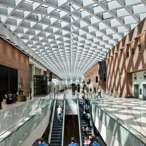 A modern, sunlit indoor space with a glass-paneled roof creating a geometric pattern. People traverse multiple floors using escalators. The area includes sitting spaces, shops, and a brick wall with diamond-shaped shadows cast by the roof design, embodying the spirit of emerging technology.