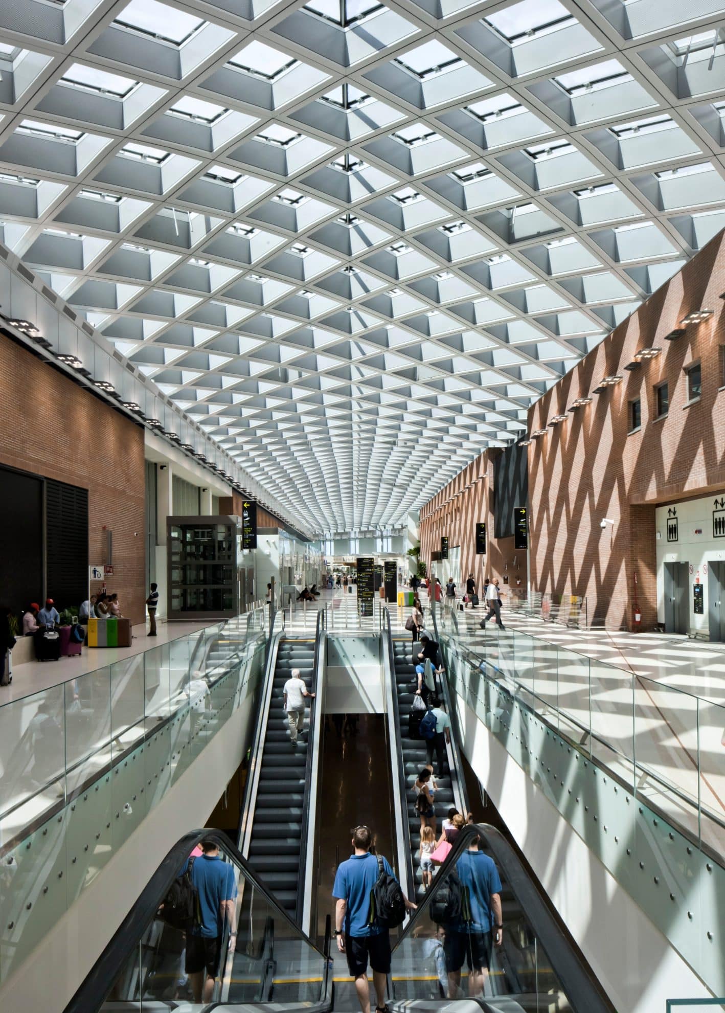 A modern, sunlit indoor space with a glass-paneled roof creating a geometric pattern. People traverse multiple floors using escalators. The area includes sitting spaces, shops, and a brick wall with diamond-shaped shadows cast by the roof design, embodying the spirit of emerging technology.