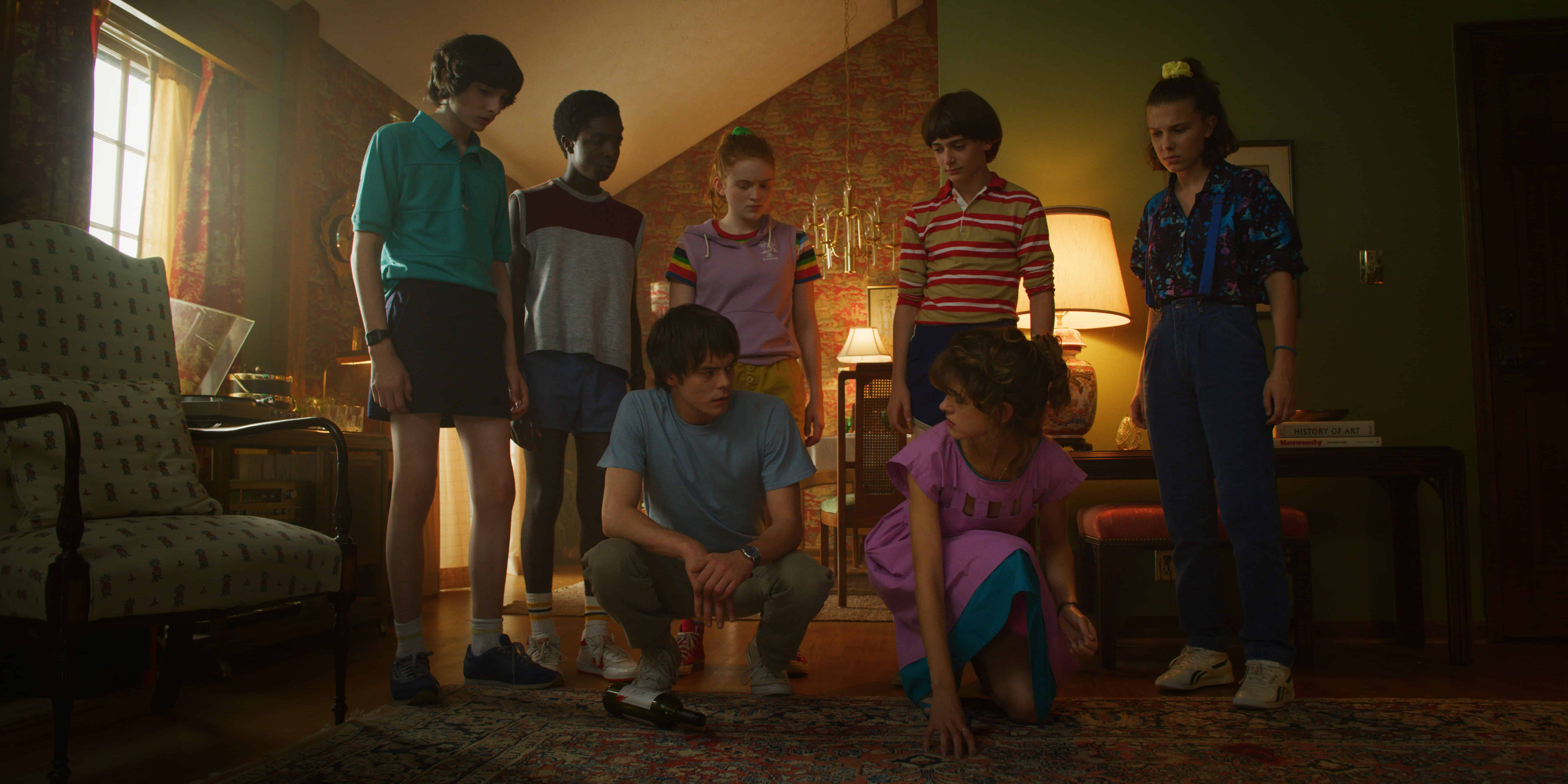 A group of seven people, some standing and some kneeling, are gathered in a dimly-lit room with vintage decor and a patterned rug. One person is crouched on the floor, examining something, while the others look on attentively, as if waiting for a Netflix update in a scene filled with quiet anticipation.