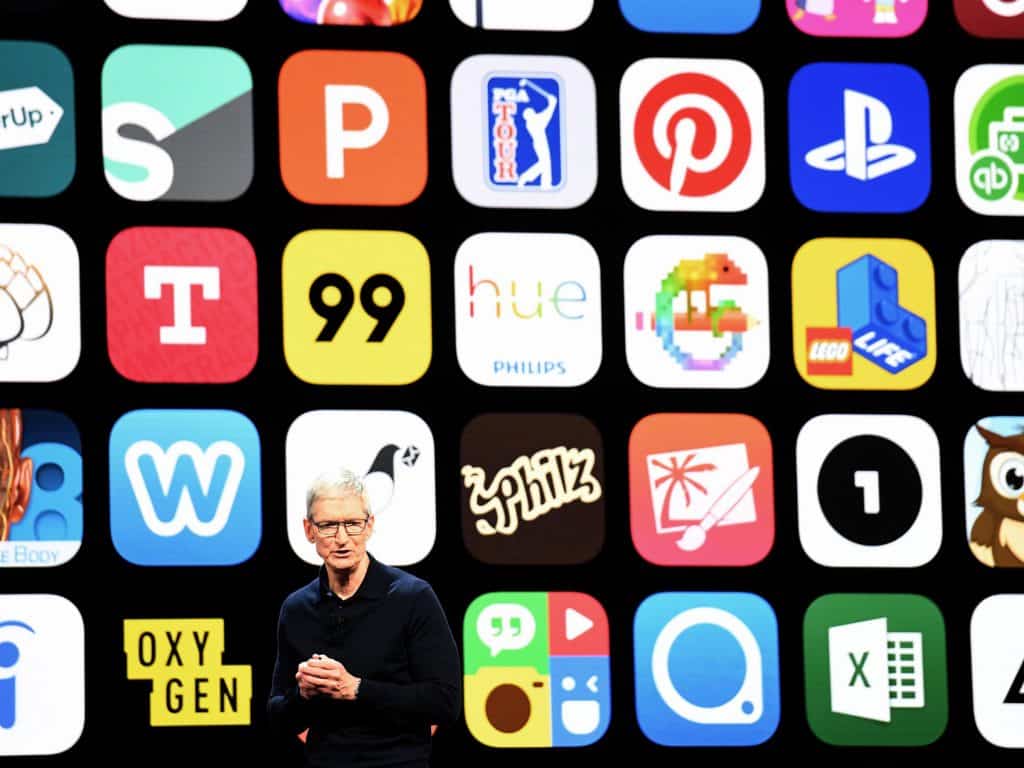 A person stands on stage in front of a large screen displaying a grid of various app icons, including popular apps like Pinterest, PlayStation, Lego, Hue, and others. The individual is wearing glasses and a black long-sleeve shirt while discussing how to top up your account using Apple ID.